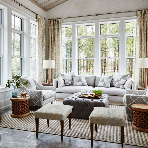 Full view of sunroom featuring a light grey couch, darker grey patterned chairs, fur topped stools, and wooden accents
